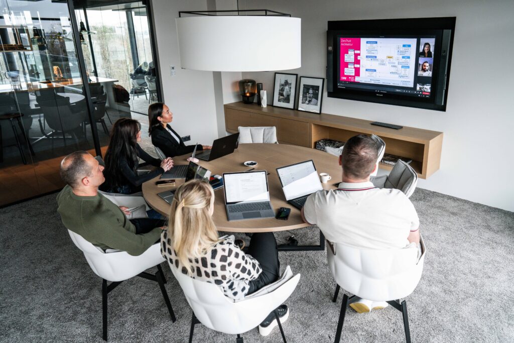 Ein Projektteam mit fünf Kollegen vor einem Bildschirm in einem Meeting mit weiteren Kollegen.